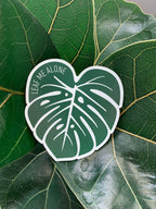 A magnet shaped like a Monstera leaf with the text 'LEAF ME ALONE' in the center, placed on a green leaf.