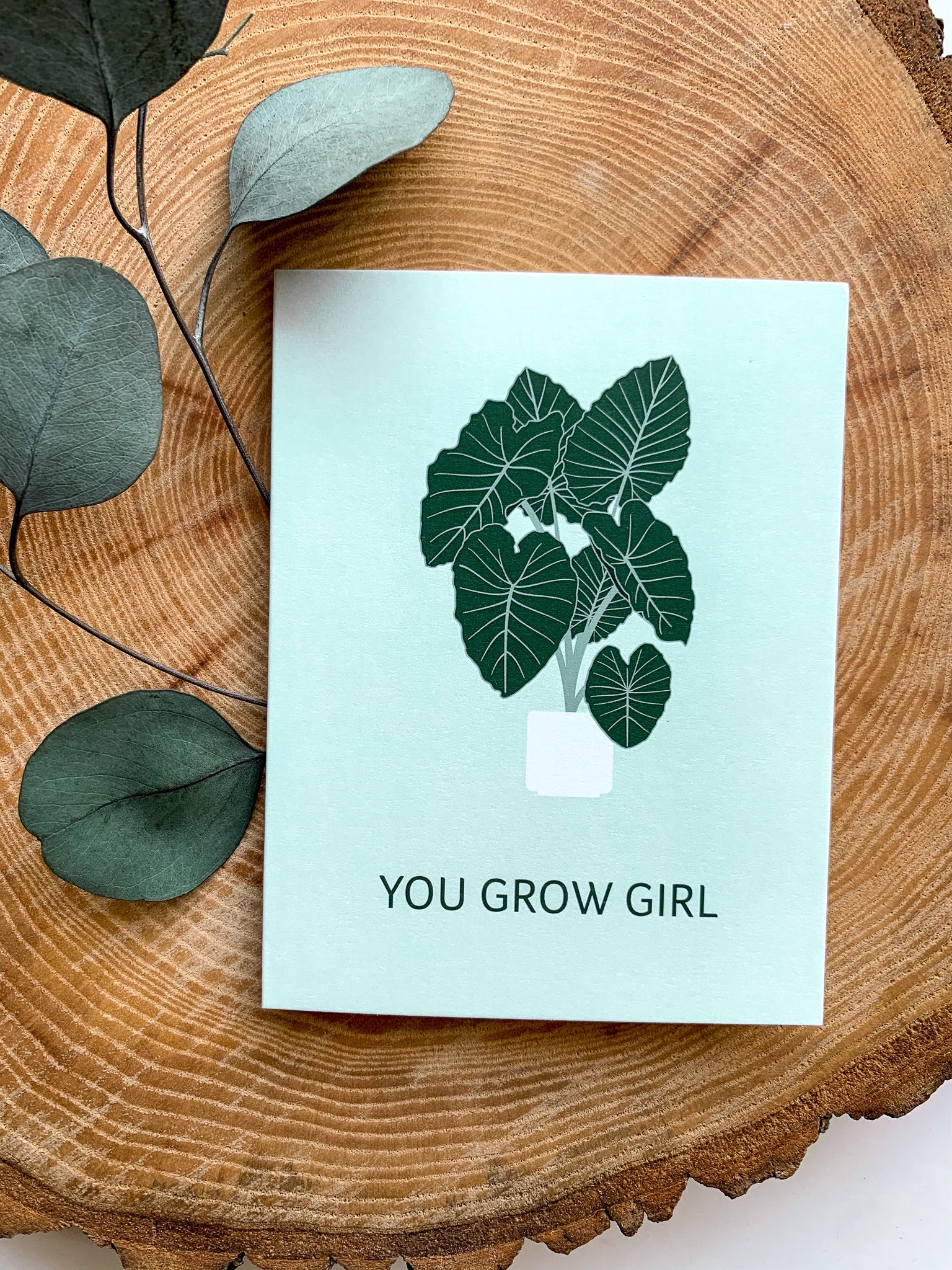 A greeting card with a plant illustration and the text 'YOU GROW GIRL' on it, placed on a wooden surface with some leaves beside it.