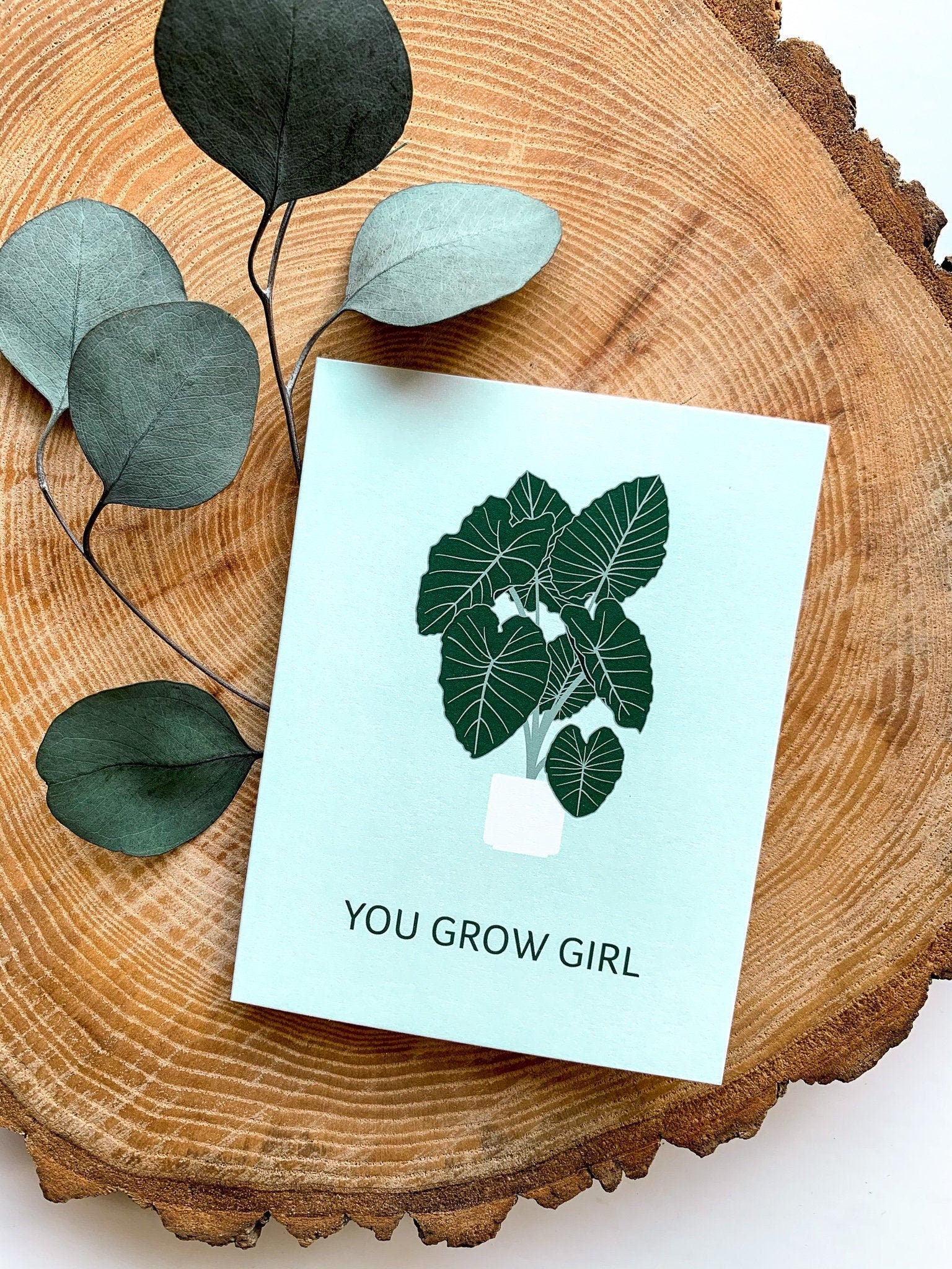 A greeting card with a plant illustration and the text 'YOU GROW GIRL' on it, placed on a wooden surface with some leaves beside it.