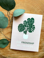 A greeting card with a plant illustration and the text 'OUR FRIENDSHIP WILL NEVER DIE' on it, placed on a wooden surface with some leaves beside it. A white envelope and plastic sleeve are also visible.