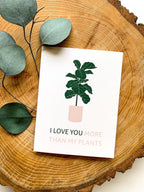 A greeting card with an illustration of a potted plant and the phrase 'I LOVE YOU MORE THAN MY PLANTS' placed on a wooden surface, accompanied by a white envelope .