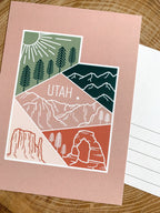A postcard with an illustrated mountain range and the text 'UTAH' prominently featured. The design includes a mix of colors representing the landscape, and a logo is visible on the bottom.