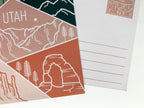 A postcard featuring a colorful illustration of Utah's landscape, including mountains and trees, with the word 'UTAH' prominently displayed.