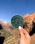 A round sticker with a green background, featuring a mountain range and the text 'Faith Moves Mountains' held against a backdrop of mountains.