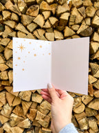 A hand holding a Christmas card with the text 'Merry & Bright' displayed, adorned with a illustrated bow and greenery, against a backdrop of autumn trees.