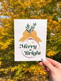 A hand holding a Christmas card with the text 'Merry & Bright' displayed, adorned with a illustrated bow and greenery, against a backdrop of autumn trees.