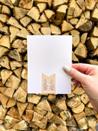 A hand holding a Christmas card with the text 'Merry & Bright' displayed, adorned with a illustrated bow and greenery, against a backdrop of autumn trees.