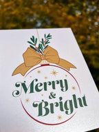 A hand holding a Christmas card with the text 'Merry & Bright' displayed, adorned with a illustrated bow and greenery, against a backdrop of autumn trees.
