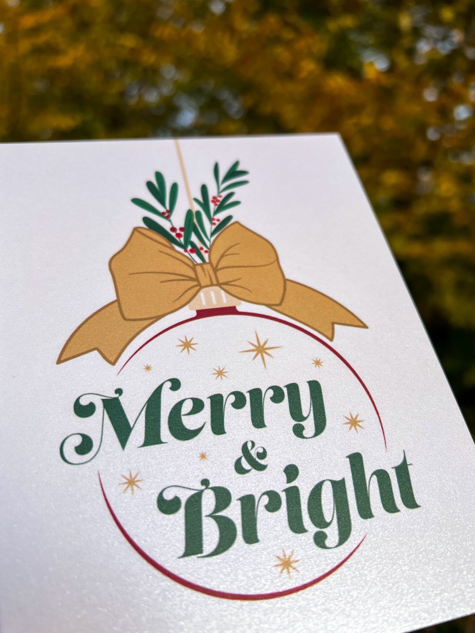 A hand holding a Christmas card with the text 'Merry & Bright' displayed, adorned with a illustrated bow and greenery, against a backdrop of autumn trees.