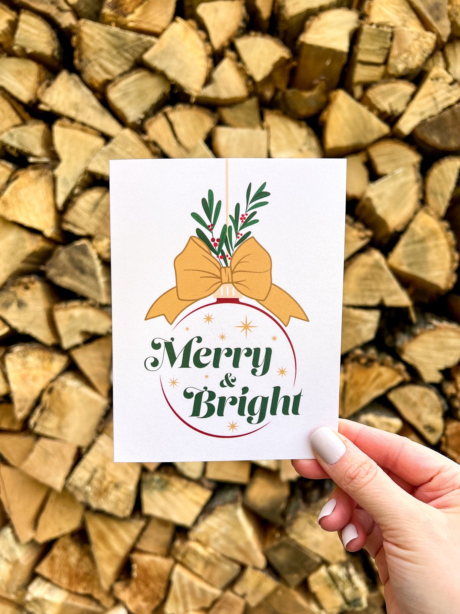 A hand holding a Christmas card with the text 'Merry & Bright' displayed, adorned with a illustrated bow and greenery, against a backdrop of autumn trees.