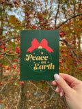A hand holding a Christmas card with 'Peace on Earth' written on it, featuring a green background, a red bow, and stars.