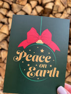 A hand holding a Christmas card with 'Peace on Earth' written on it, featuring a green background, a red bow, and stars.