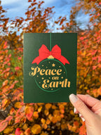 A hand holding a Christmas card with 'Peace on Earth' written on it, featuring a green background, a red bow, and stars.