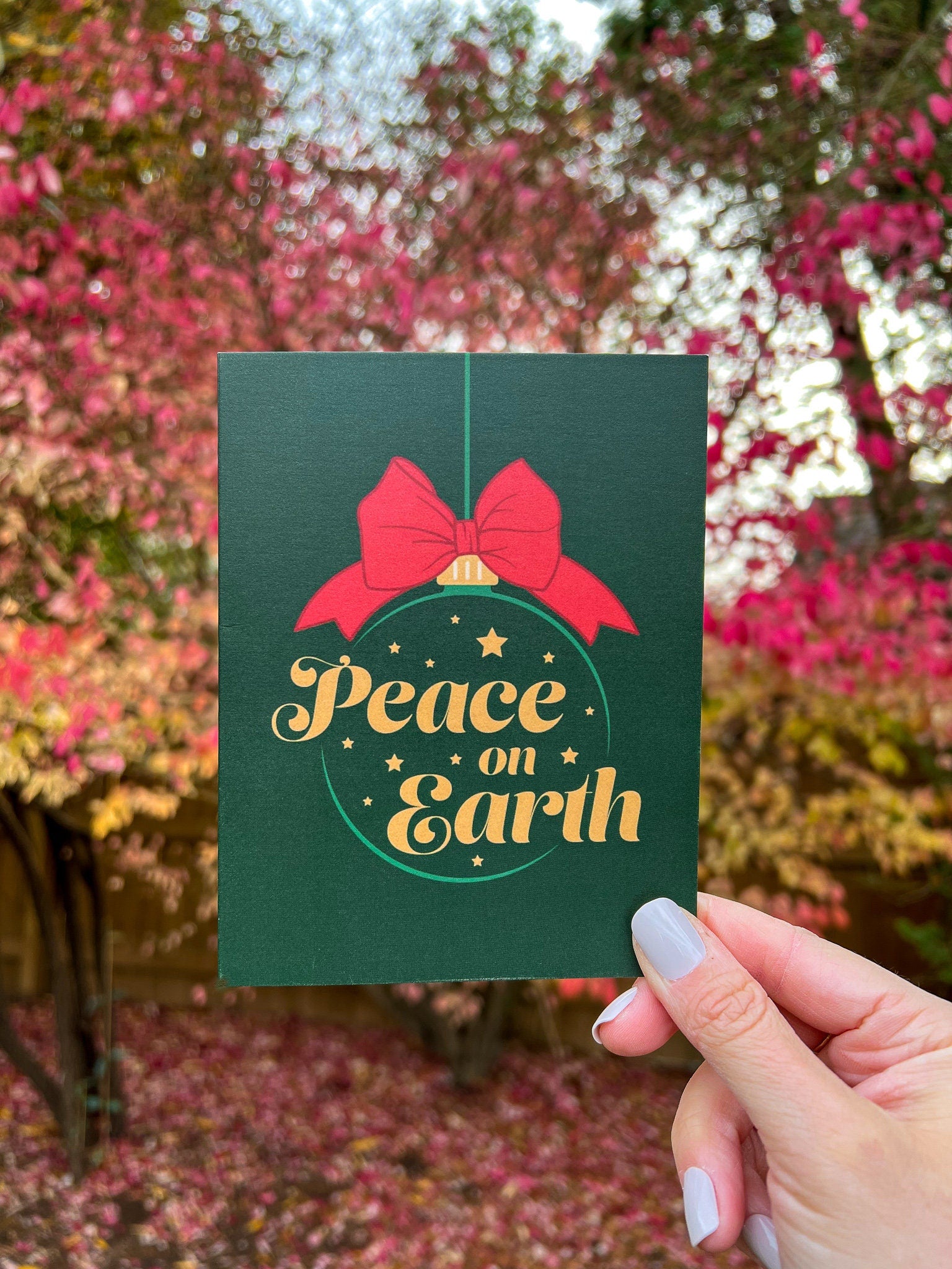 A hand holding a Christmas card with 'Peace on Earth' written on it, featuring a green background, a red bow, and stars.
