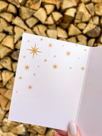 A hand holding a Christmas card with the text 'Merry & Bright' displayed, adorned with a illustrated bow and greenery, against a backdrop of autumn trees.