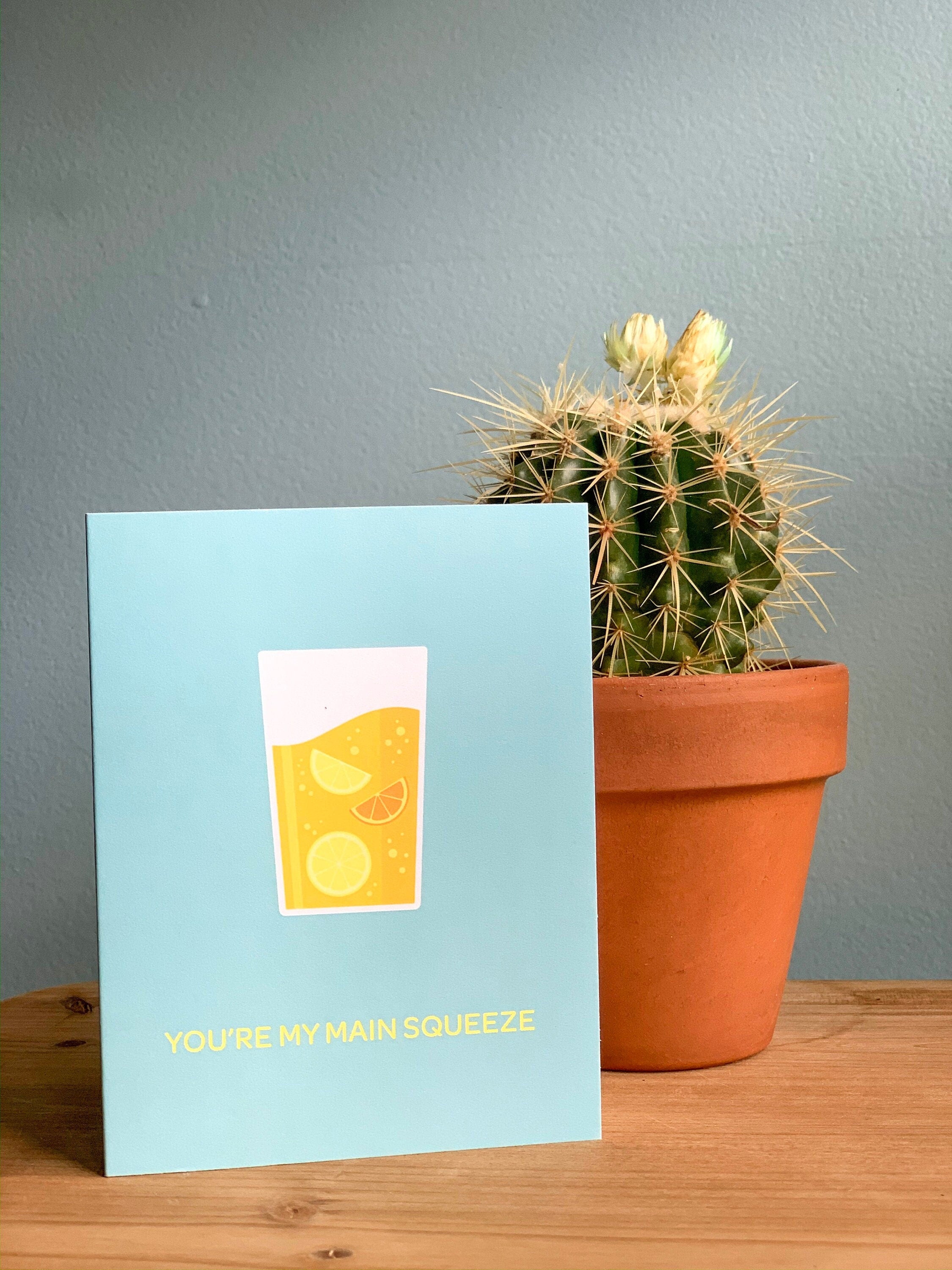 A greeting card with an illustration of a lemonade in a glass, placed on a wooden surface with a cactus in the background.