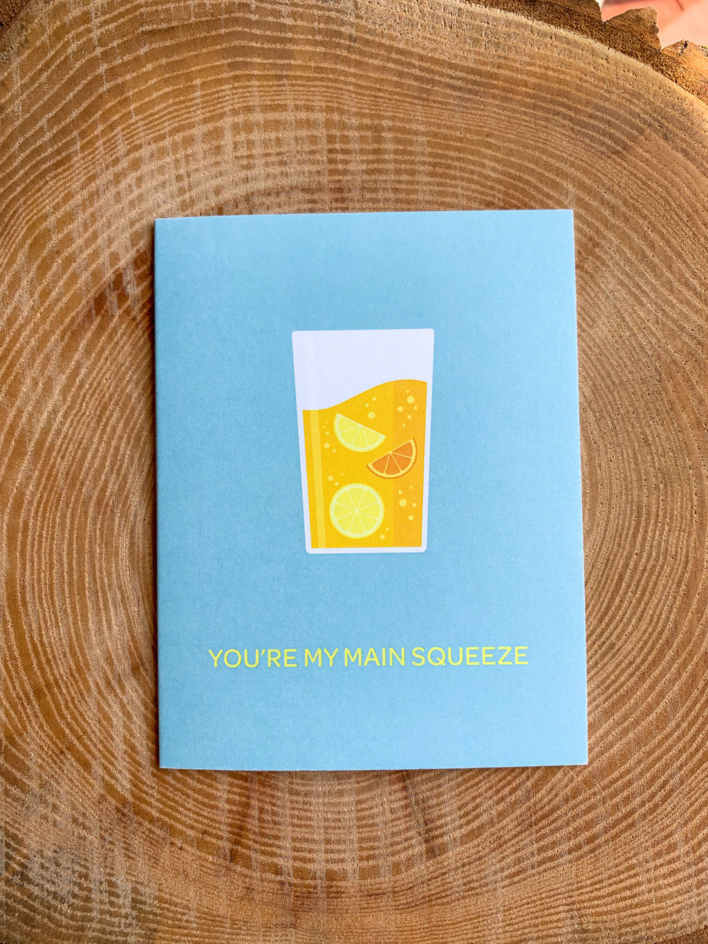 A greeting card with an illustration of a lemonade in a glass, placed on a wooden surface with a cactus in the background.