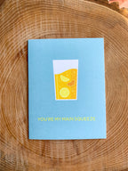 A greeting card with an illustration of a lemonade in a glass, placed on a wooden surface with a cactus in the background.