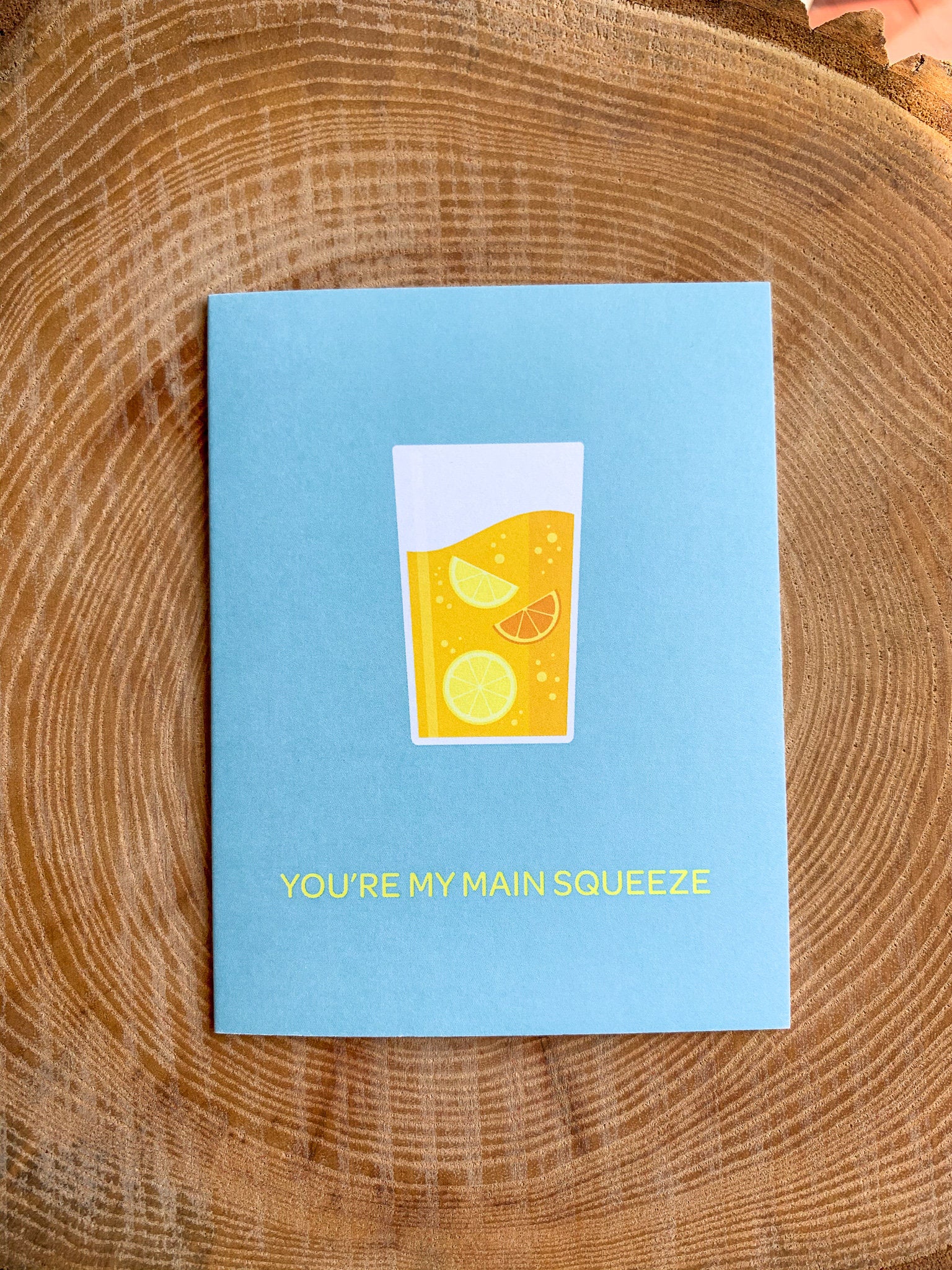 A greeting card with an illustration of a lemonade in a glass, placed on a wooden surface with a cactus in the background.