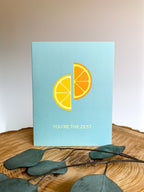 A greeting card with an illustration of a lemon and an orange, with the pun 'YOU'RE THE ZEST' printed on the front, placed on a wooden surface with a white envelope and green leaves in the background.