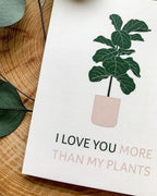 A greeting card with an illustration of a potted plant and the phrase 'I LOVE YOU MORE THAN MY PLANTS' placed on a wooden surface, accompanied by a white envelope .