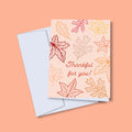 A thank you greeting card with leaf patterns and the text 'Thankful for you!' on it, accompanied by a white envelope, displayed against an orange background.